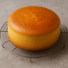 Génoise dorée et moelleuse, fraîchement sortie du four, posée sur une grille de refroidissement. Sa surface lisse et légèrement bombée reflète la lumière naturelle d’une cuisine chaleureuse, évoquant une texture aérienne et fondante.