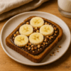 Toast au pain complet garni de chocolat noir fondu, de rondelles de banane et d’éclats de noisettes, présenté sur une assiette en céramique claire avec un fond en bois.