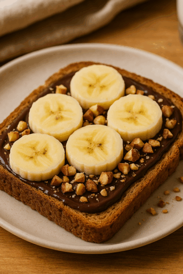 Toast au pain complet garni de chocolat noir fondu, de rondelles de banane et d’éclats de noisettes, présenté sur une assiette en céramique claire avec un fond en bois.