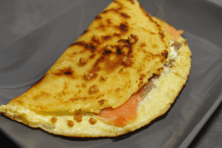 Brique au saumon garnie de crème fraîche et de petits éclats de noix, servie chaude avec un léger filet de fumée sur une assiette grise.