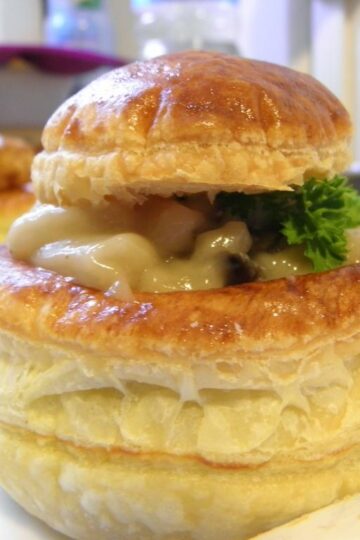 Vol-au-vent feuilleté doré, garni d’une crème onctueuse aux champignons. Le chapeau du feuilleté est légèrement posé de travers, laissant apparaître la garniture crémeuse et un brin de persil frais.