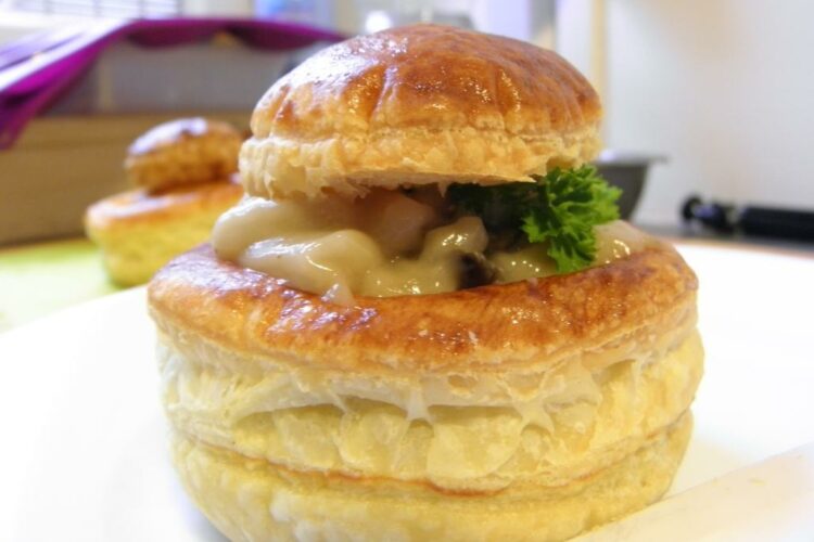 Vol-au-vent feuilleté doré, garni d’une crème onctueuse aux champignons. Le chapeau du feuilleté est légèrement posé de travers, laissant apparaître la garniture crémeuse et un brin de persil frais.