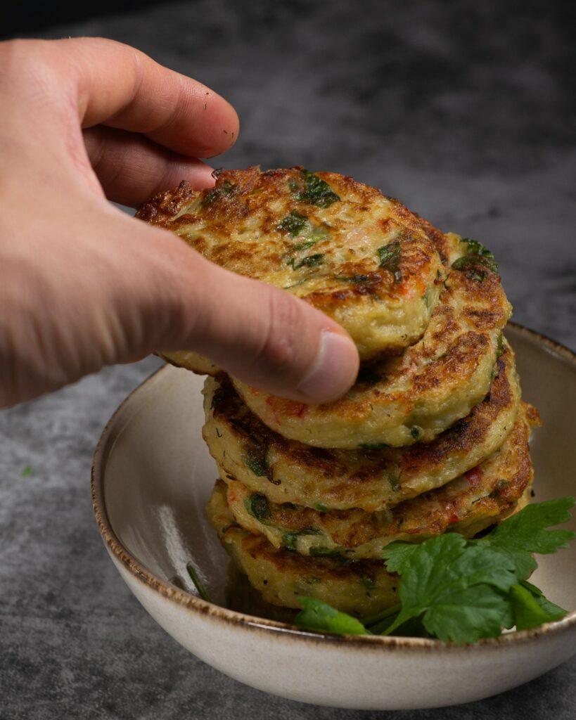 Beignets de courgettes dorés et moelleux, empilés dans un petit bol, avec une main qui en saisit un au-dessus. Les galettes sont bien grillées, parsemées d’herbes fraîches, sur un fond gris texturé.