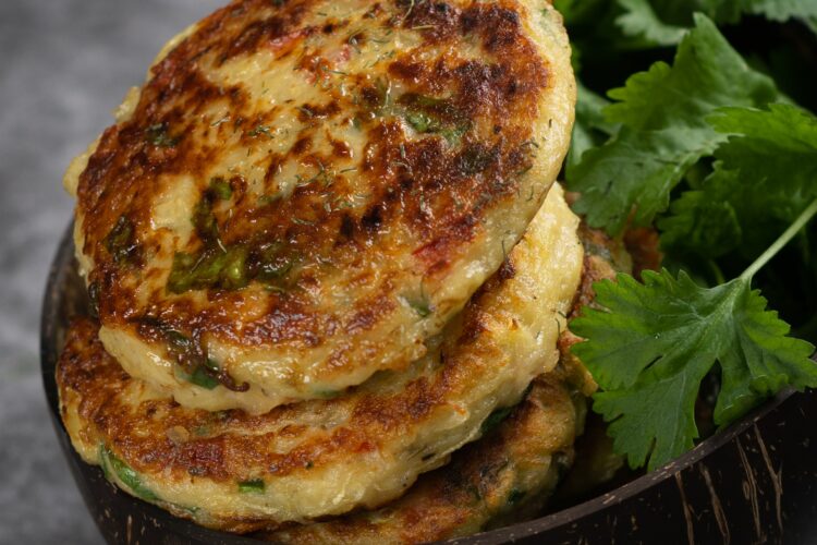 Beignets de courgettes dorés et moelleux, empilés dans un petit bol, avec une main qui en saisit un au-dessus. Les galettes sont bien grillées, parsemées d’herbes fraîches, sur un fond gris texturé.