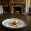 Une assiette d’hiver raffinée posée sur une table en bois foncé, illuminée par la lueur d’un feu de cheminée. Au centre, de délicates étoiles dorées renfermant un cœur de châtaigne, entourées de pétales colorés et d’une mousse de poire safranée, dans une atmosphère chaleureuse et festive de décembre.