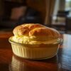 Soufflé doré et moelleux dans un plat en verre, posé sur une table en bois foncé dans un salon baigné de lumière naturelle.